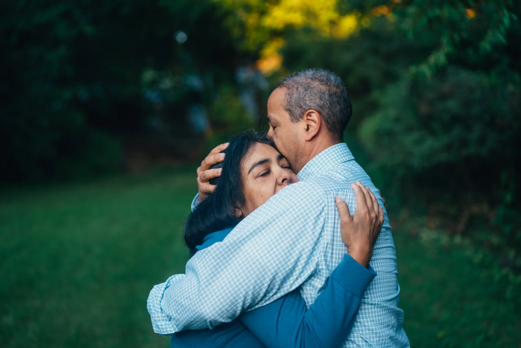 Two adults sharing a warm embrace outdoors, one kissing the other's forehead